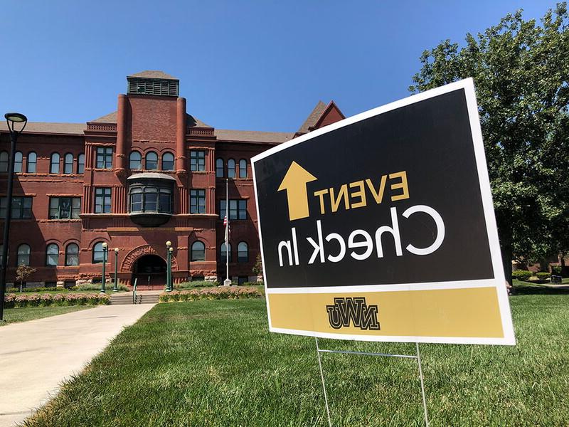 A sign staked into the ground in front of the the Old Main building reads: Event check-in with and up arrow. 