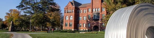 A sculpture shaped in the letter O, in front of the large red-brick building of Old Main.